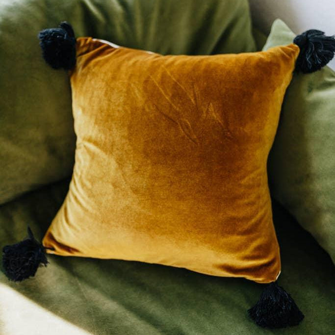A luxurious gold velvet pillow with black tassels on a cream canvas back, placed on a green sofa.